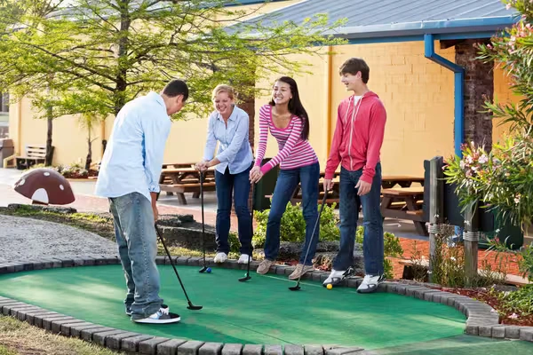 Padre jugando mini golf mientras su esposa e hijos observan y ríen, esperando su turno en un ambiente familiar y divertido.