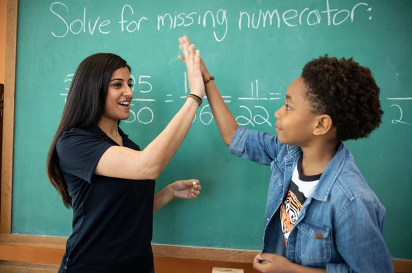 Niño y profesor frente a una pizarra con ecuaciones matemáticas, celebrando con un choque de manos tras comprender el ejercicio.