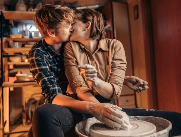 Pareja besándose junto a un torno de cerámica en un taller de arte, compartiendo un momento romántico y creativo.