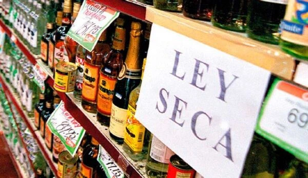 Estanterías de una tienda de licores con botellas alineadas y un gran cartel que dice “Ley Seca”, indicando que no se puede comprar alcohol ese día.