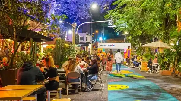 Calle peatonal junto a Parque Lleras en Medellín con personas sentadas en mesas al aire libre, conversando y riendo en bares y restaurantes.