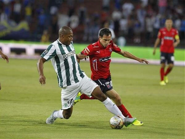 Partido de fútbol en Medellín entre los dos equipos locales, con dos jugadores rivales disputando el balón mientras otros futbolistas observan la jugada en el campo.