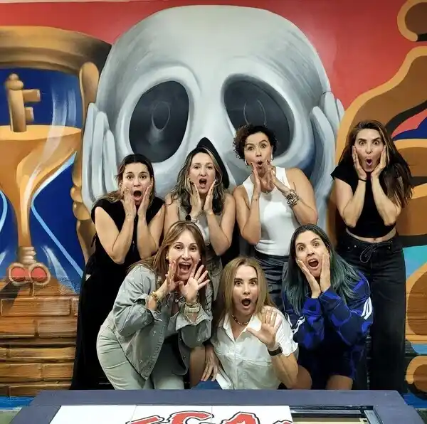 Siete mujeres posan frente a un mural de grafiti inspirado en El Grito de Edvard Munch, con las manos en las mejillas imitando la expresión icónica de la obra.