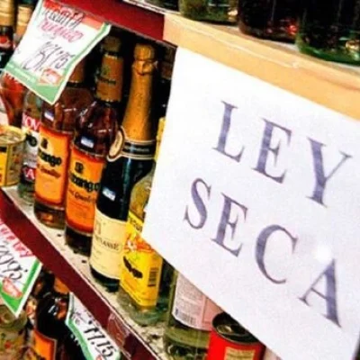 Estanterías de una tienda de licores con botellas alineadas y un gran cartel que dice “Ley Seca”, indicando que no se puede comprar alcohol ese día.