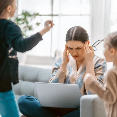 Madre trabajando en un computador mientras sus hijos gritan y juegan alrededor, mostrando expresión de estrés y cansancio.