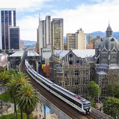 Panorámica de la ciudad de Medellín con el Metro pasando por el centro, rodeado de edificios modernos y montañas verdes al fondo.